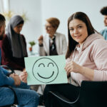 Young happy woman holding holding placard with drawn smiley face during group therapy meeting at mental health center.