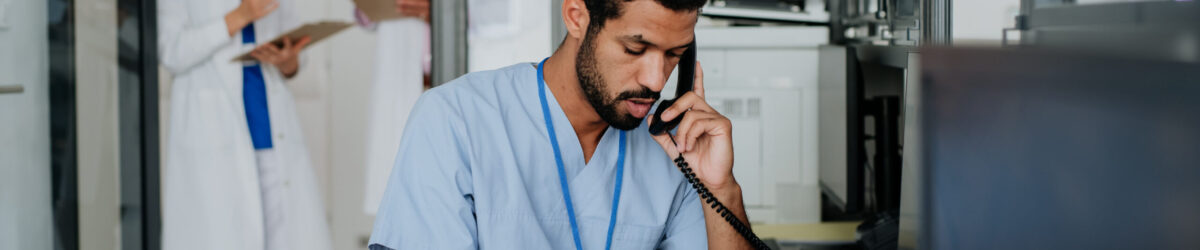 Young multiracial nurse sitting at reception and calling,his colleagues discussing in the background.