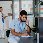 Young multiracial nurse sitting at reception and calling,his colleagues discussing in the background.